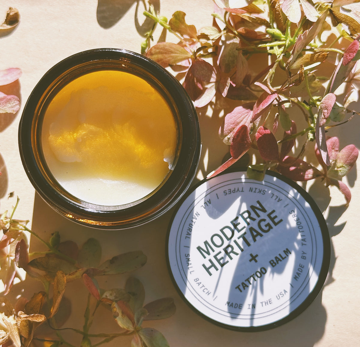 Open jar of Modern Heritage tattoo balm beside green foliage in warm light